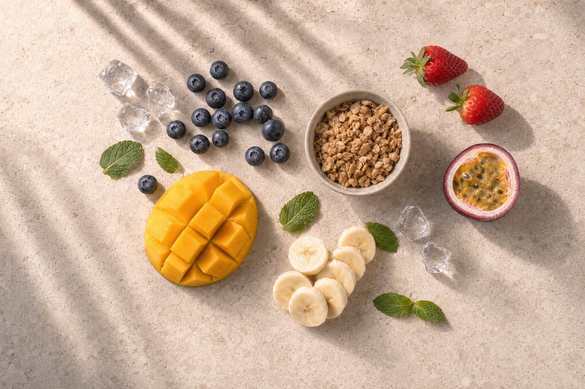 Mango, blueberries, banana, strawberries, passion fruit, granola and mint laid out on stone